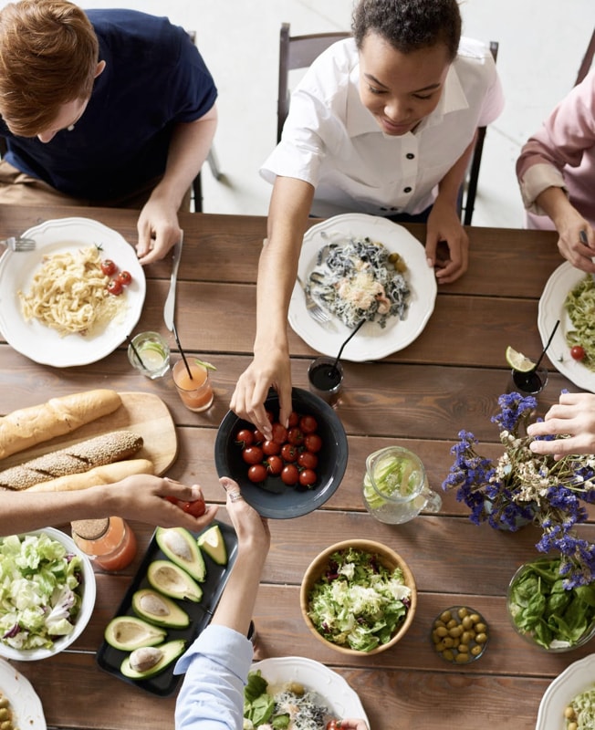 Group of people sharing a meal, with plates of pasta, salads, sliced avocados and cherry tomatoes on a wooden table.