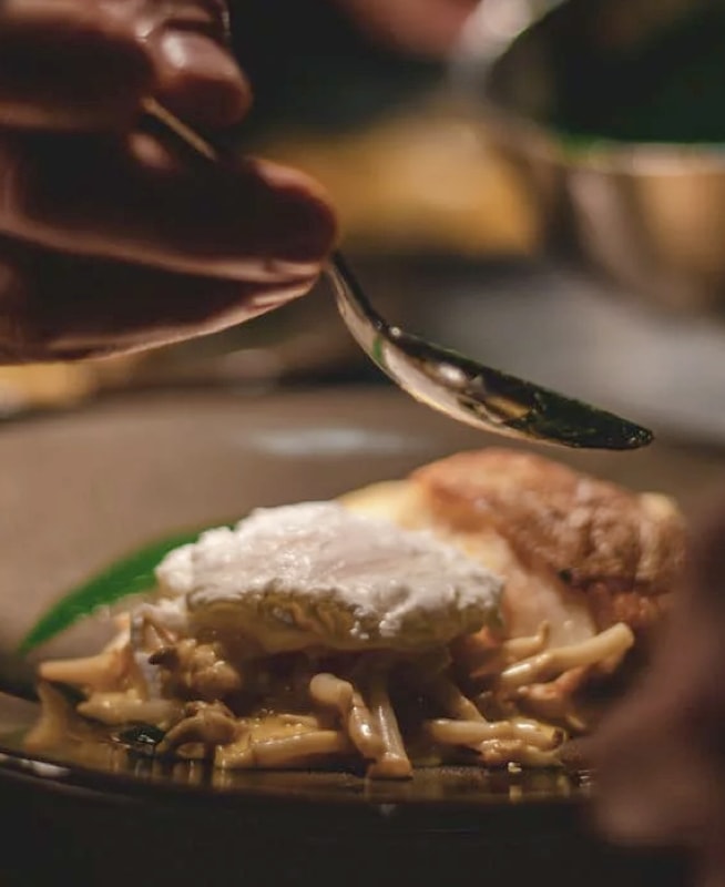 Chef's hands holding a spoon over a plate of plated dish with pasta, white sauce and a garnish.